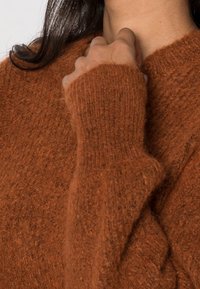 Pull marron en maille avec une texture duveteuse, une coupe ample et des manches longues avec des poignets côtelés. Les détails du tissu sont visibles.