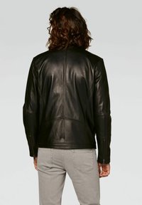 Giacca di pelle nera con un design aderente, dettagli delle cuciture notevoli e polsini con zip. Indossata sopra dei jeans grigi, vista da dietro.