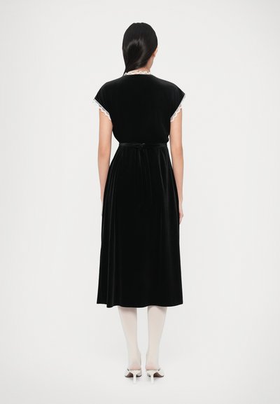 Vestido de veludo preto com mangas curtas tipo capa, acabamento em renda e cinto na cintura. O vestido tem comprimento até ao meio da perna, com uma textura suave e macia.