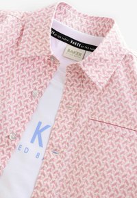 Růžová košile s krátkým rukávem a geometrickým vzorem, bílým prošíváním. Obsahuje límeček na knoflíky a kapsu na hrudi, vrstvenou přes bílý tričko.