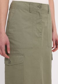 Gros plan d'une personne portant une jupe cargo vert olive avec poche à rabat boutonnée, poches latérales et passants pour ceinture.