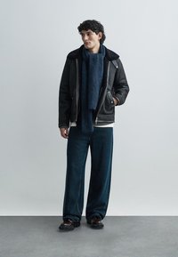 Veste en cuir noir avec col en peau lainée, écharpe texturée bleu marine, chemise blanche, pantalon en denim foncé ample, et chaussures noires épaisses.