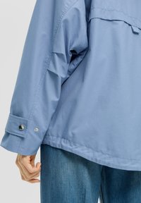 Veste surdimensionnée bleu clair avec des manches longues, un détail de bouton-pression au niveau du poignet et une texture lisse. Portée par-dessus un tissu en denim.