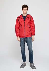 Rød vandtæt jakke med hætte og snore, med marineblå detaljer og to forlommer. Båret over en marineblå skjorte og blå jeans.