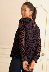 Blusa nera e viola a righe zebra con maniche trasparenti, scollatura alta e polsini con bottoni. Presenta un'apertura a chiave sul retro.