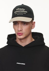 Crna bejzbol kapa s svijetlosmeštim vizirama, sa natpisom "sportska udruga". Nosite s crnom duksericom koja prikazuje bijeli logo.
