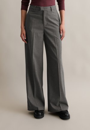 Grau gestreifte, weit geschnittene Hose mit taillierter Form, die eine hohe Taille und eine glatte Stofftextur aufweist. Kombiniert mit klobigen schwarzen Schuhen.
