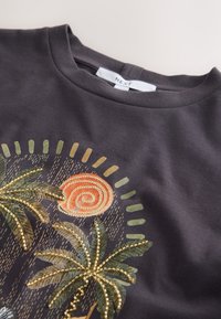 T-shirt gris foncé avec des palmiers brodés et un motif de soleil en spirale accentué de perles dorées, étiquette "NEXT" au col.