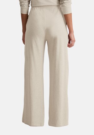 Person, die beige weit geschnittene Hosen trägt, von hinten gezeigt, dazu ein passendes langärmliges Oberteil vor einem schlichten Hintergrund.