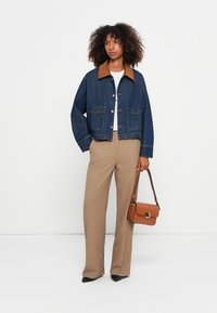Vrouw met krullend haar, gekleed in een blauwe spijkerjack, beige wijde broek, zwarte schoenen en een bruine handtas vast.