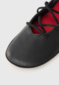 Zwarte leren schoen met een gestructureerde afwerking, met een ronde neus en een rode binnenkant. Zwarte veters kruisen over een uitsnijdontwerp bovenop.