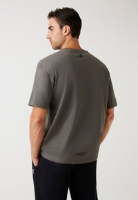 Hombre con cabello oscuro que lleva una camiseta de manga corta gris y pantalones negros, de pie con las manos en los bolsillos, de espaldas frente a un fondo plano.