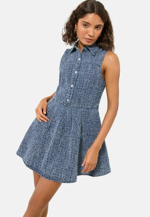 Frau trägt ärmelloses, strukturiertes blaues Hemdkleid mit Kragen, steht mit der Hand in der Hüfte vor schlichtem Hintergrund.