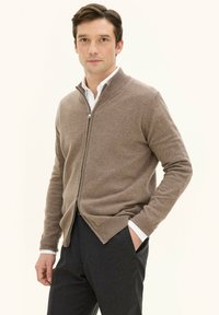 Maglione marrone con zip e colletto alto, realizzato in un tessuto morbido, abbinato a una camicia bianca e pantaloni scuri. Design semplice senza fantasie.