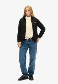 Veste en polaire noire avec un col montant, sous un pull en tricot crème, associée à un jean bleu baggy et des chaussures noires.