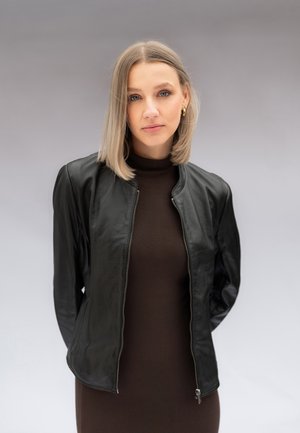 Junge Frau mit blondem Haar, die goldene Creolenohrringe trägt, eine schwarze Lederjacke über einem dunkelbraunen Oberteil, stehend vor einem grauen Hintergrund.