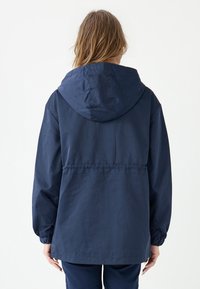 Giacca a cappuccio blu navy con polsini elasticizzati, vita cintrata e vestibilità rilassata. Tessuto liscio e leggero con finitura opaca.