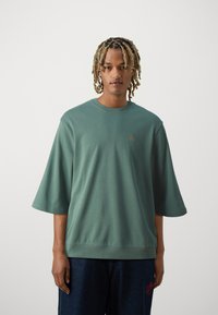 Grøn oversized T-shirt med korte ærmer, rund halsudskæring, tekstureret stof og små broderede detaljer på brystet. Bærer den med mørke denim.