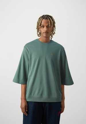 Green oversized T-shirt with short sleeves, round neckline, textured fabric, and small embroidered detail on the chest. Worn with dark denim.