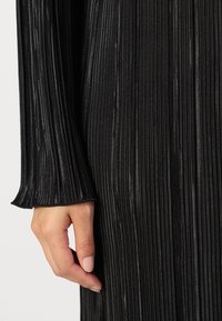 Μαύρο πλισέ φόρεμα με μανίκι και υφασμάτινη υφή και φαρδιά μανσέτα, που δείχνει λεία επιφάνεια και απαλή γυαλάδα. Χέρι τοποθετημένο δίπλα.