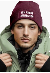Maroon knit beanie with the text "ICH HASSE MENSCHEN," worn with a light green, padded jacket, featuring a black inner layer and visible buttons.