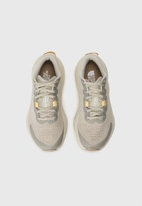 Des chaussures de sport beige clair avec une tige en mesh, des accents dorés et des lacets gris. Des surfaces texturées et une forme arrondie améliorent le confort et la stabilité.