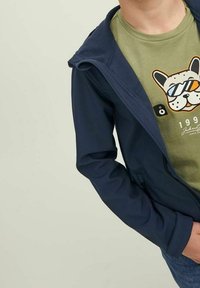 Child wearing a green t-shirt with a dog wearing sunglasses graphic, layered under an unzipped navy blue jacket.