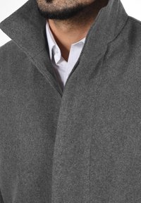 Abrigo de lana gris con un cuello alto y un acabado texturizado limpio. Presenta un cuello abotonado y un detalle de costura. Camisa blanca visible debajo.
