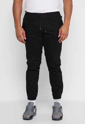 Homme portant un pantalon de jogging noir avec des poignets élastiques, des chaussettes blanches, des baskets Nike grises et bleues, et une chemise blanche sur un fond uni.