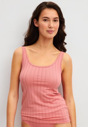 Femme aux longs cheveux foncés portant un haut sans manches rose côtelé, debout devant un fond clair uni.