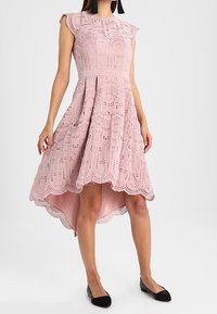 Vaaleanpunainen pitsimekko, jossa on monimutkaisia kuvioita, korkea-alamittainen helma ja hihattomat hihat. Mustien ballerinojen kanssa se esittelee leikkausdetaljien. 