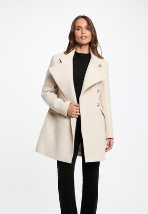 Vrouw draagt een beige dubbelgeknoopte jas over een zwarte trui en broek, staand met één hand de jas vasthoudend.
