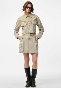 Veste en denim beige coupée, avec deux poches sur la poitrine, associée à une jupe en denim assortie avec des poches latérales et un ourlet effiloché, portée avec des bottes noires.