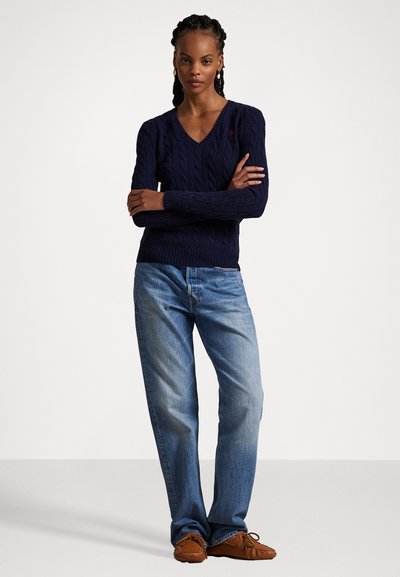 Tamsiai mėlynas megztinis su kabelių raštu ir V formos iškirpte, derinamas su šviesiai mėlynomis džinsomis ir rudiems mokasininio tipo batams. Paprastas dizainas ir tekstūruota medžiaga.