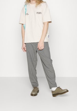 Beige bomulls t-shirt med tryckt text, kombinerad med grå byxor med vertikala ränder. Fotbeklädnad: bruna läderskor med rund tå.