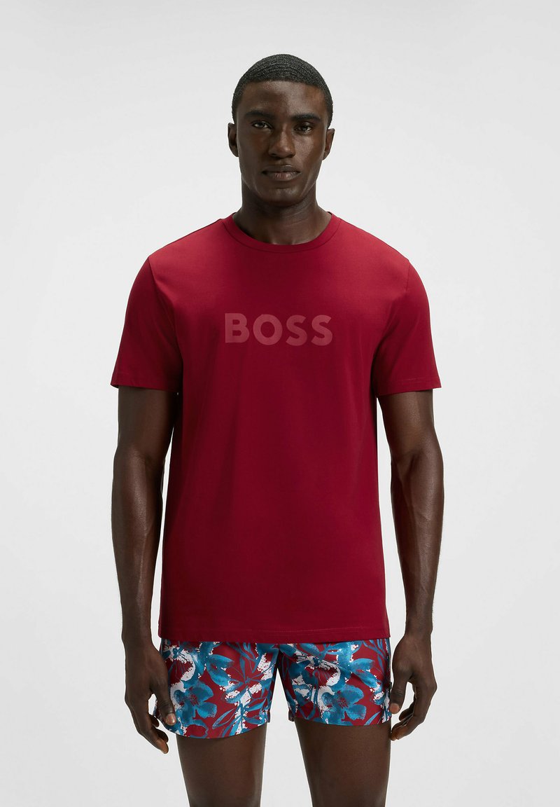 Rotes T-Shirt mit Rundhalsausschnitt und dezentem "BOSS"-Logo, kombiniert mit blauen, blumigen Badeshorts mit weißen Akzenten und einer sanften Stofftextur.