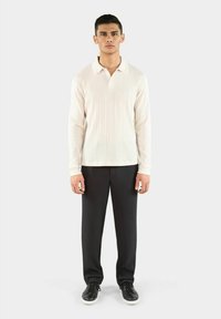 Langärmliges cremefarbenes Poloshirt mit gerippter Textur, kombiniert mit schwarzen Nadelstreifenhosen und schwarzen Sneakers, stehend vor einem neutralen Hintergrund.