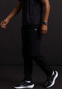 Zwarte sportbroek met een slim fit, met een zichtbaar logo, een elastische tailleband en zakken. Gepaard met zwarte sneakers en een horloge.