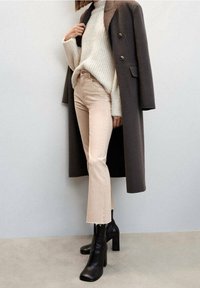 Jean cropped beige, pull crème épais, et un long manteau marron foncé associé à des bottes à talons noires. Ourlet effiloché sur les jeans.