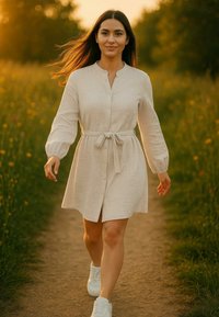 Abito button-up beige chiaro con maniche lunghe e dettaglio in vita con cintura, abbinato a sneakers bianche. Ambiente naturale con un sentiero erboso.
