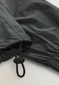 Țesătură cu carouri negre, cu un model în grilă, care prezintă un șnur reglabil și un detaliu strâns la manșetă. Ușoară și texturată.