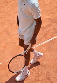 Jasnoniebieska koszulka polo tenisowa i spodenki, trzymający czarną rakietę tenisową z białym uchwytem, noszący białe skarpetki i jasne trampki, stojący na korcie ziemnym.