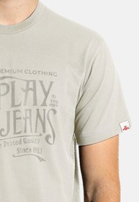 Světle šedé bavlněné tričko s kulatým výstřihem, s vybledlým grafickým potiskem s nápisem "PLAY JEANS" a malou červenou nášivkou na rukávu.