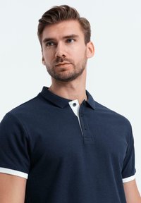 Polo azul marino con cuello y puños blancos, confeccionado en tela de algodón texturizado. Presenta una tapeta de dos botones y mangas cortas.