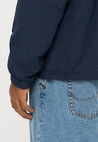 Geacă matlasată albastru marin, ușoară, cu manșete elastice, croială scurtă și tiv strâns. Blugi din denim cu șnur și textură deschisă la culoare.