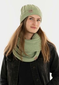 Lichtgroene gebreide beanie en bijpassende infinity sjaal met een geometrisch patroon. Beide hebben een klein label. Model draagt een zwarte jas.