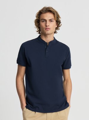 Ragazzo con capelli biondi ricci che indossa una maglietta henley blu navy a maniche corte e pantaloni beige, in piedi con le mani in tasca.
