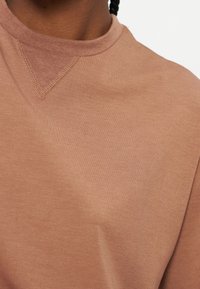 Gros plan sur une personne portant un sweat-shirt marron à col rond avec un détail de couture triangulaire visible sous le col.