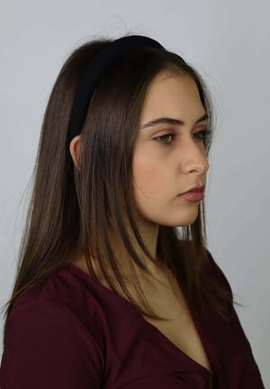 Schwarzes Stoff-Stirnband, getragen von einer Person mit langem, glattem braunem Haar, mit einer glatten Textur. Das Outfit ist burgunderfarben.