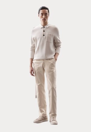 Polo de punto beige claro con cuello, tres botones oscuros y un bolsillo en el pecho. Combinado con pantalones crema claros y zapatillas beige.